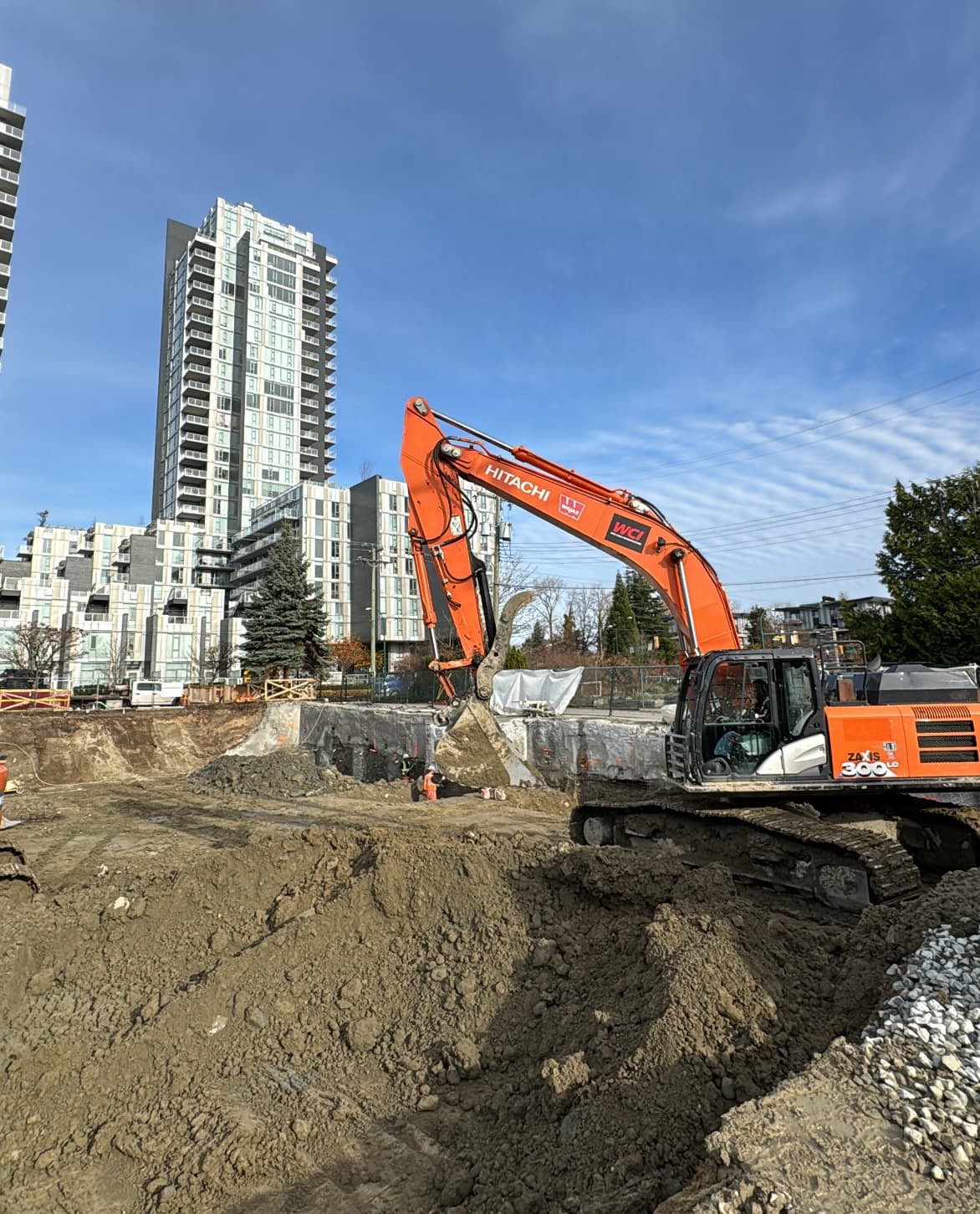 Hitachi excavator working in a deep urban excavation pit with shoring and high-rise buildings beyond.